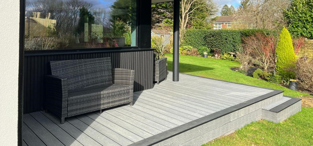 Light grey decking area bordering a black building. The decking backs onto a garden.