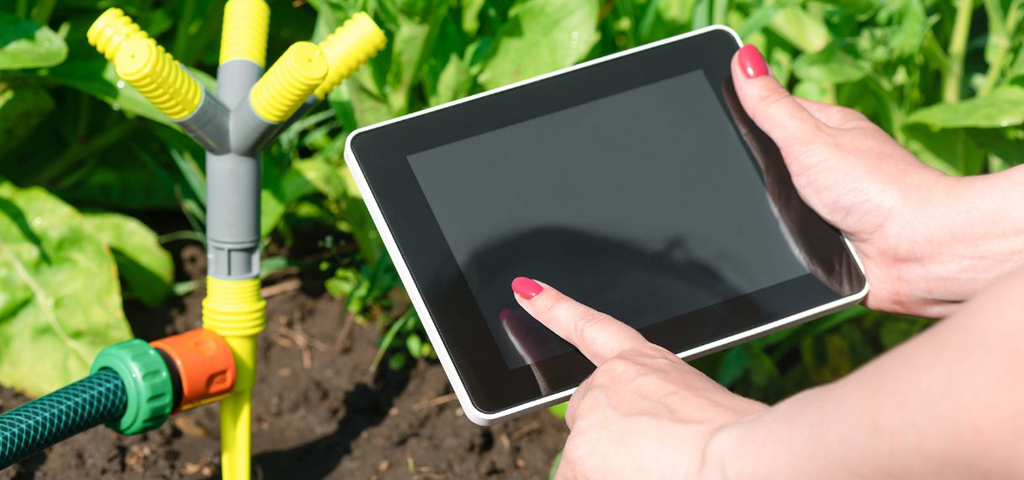 Woman's hand using a screen next to a sprinkler next to some vegetation.